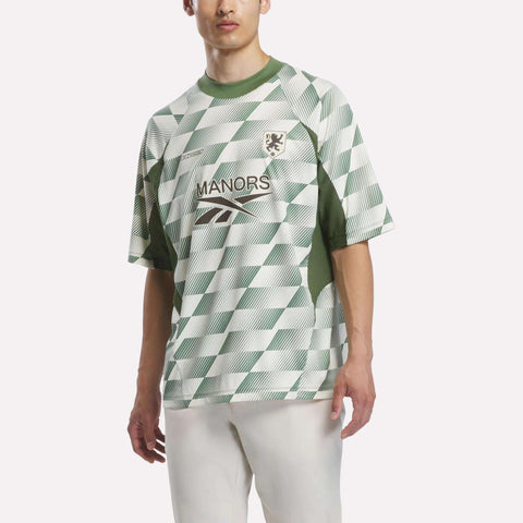 A person stands against a plain background wearing the Reebok x Manors Men’s Mockneck Polo—a green and white checkered short-sleeve jersey crafted from performance stretch fabric with a MANORS chest logo—paired with white pants.