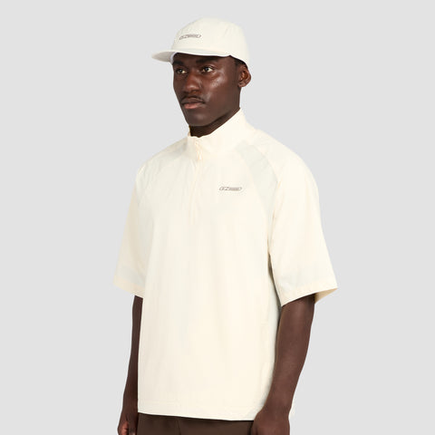 A man dressed in the Reebok x Manors Men’s Tour Shirt by Reebok, paired with a coordinating cap, stands against a plain gray background, gazing slightly to the side with a neutral expression.