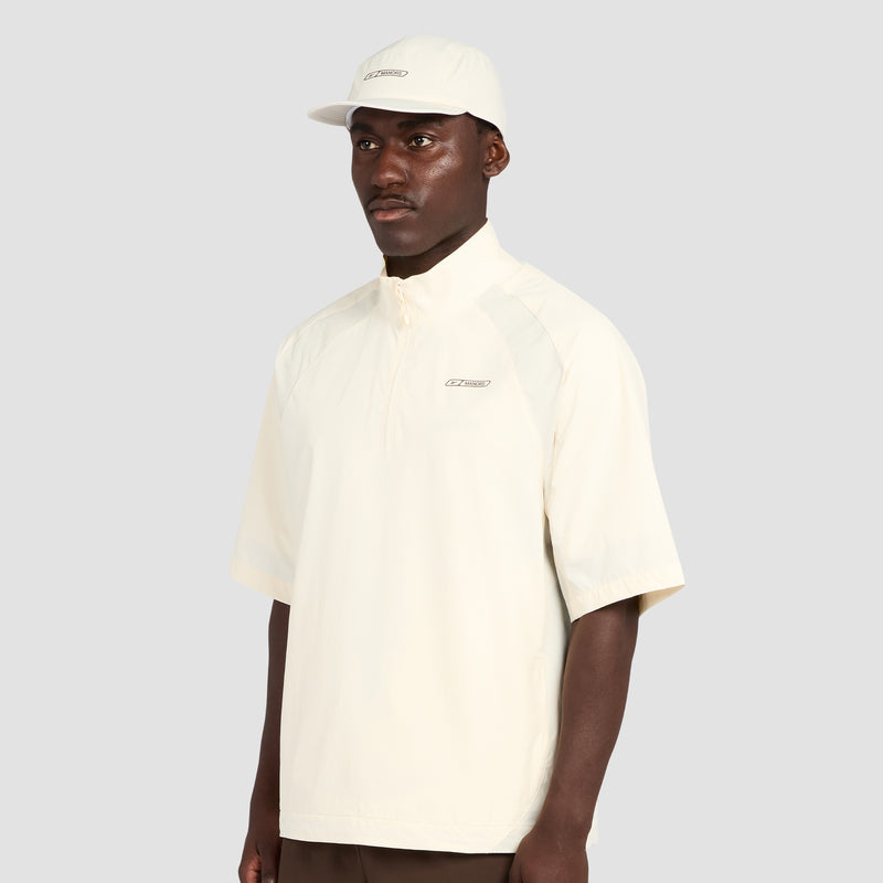 A man dressed in the Reebok x Manors Men’s Tour Shirt by Reebok, paired with a coordinating cap, stands against a plain gray background, gazing slightly to the side with a neutral expression.