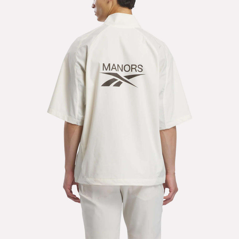 A person stands facing away, wearing the Reebok x Manors Men’s Tour Shirt—an oversized white short-sleeve golf shirt featuring MANORS and the Reebok logo printed on the back—paired with white pants, set against a plain light background.