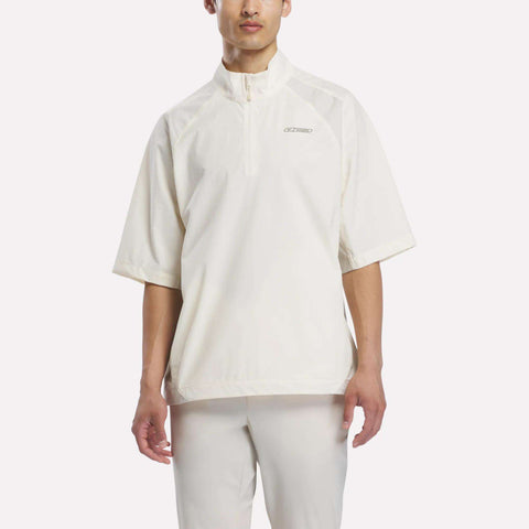 A person wearing the Reebok x Manors Men’s Tour Shirt—a loose-fitting, off-white men’s golf tour shirt with short sleeves, a half-zip collar—paired with matching pants, standing against a plain light background.