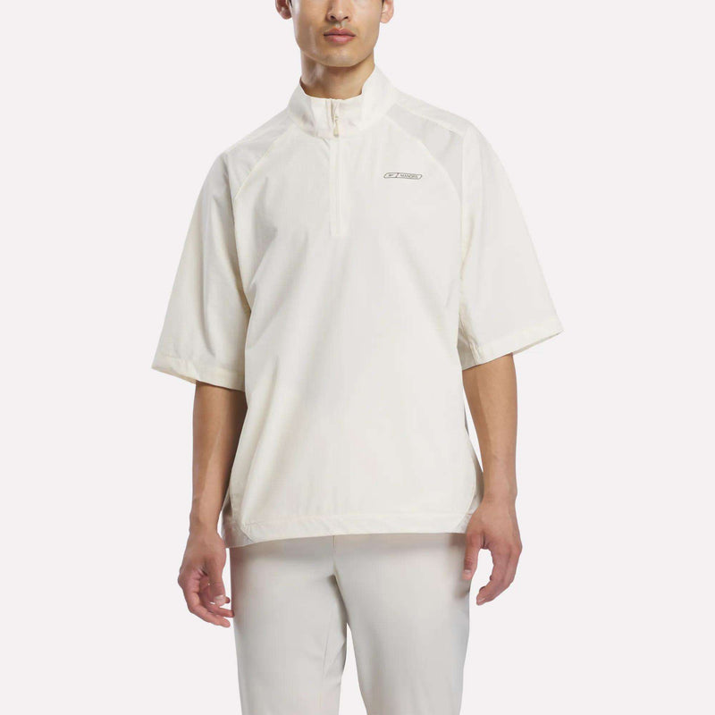 A person wearing the Reebok x Manors Men’s Tour Shirt—a loose-fitting, off-white men’s golf tour shirt with short sleeves, a half-zip collar—paired with matching pants, standing against a plain light background.