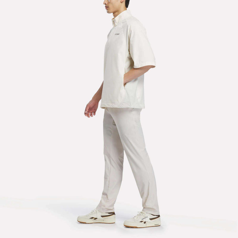 A person wearing the Reebok x Manors Men’s Tour Shirt in off-white, paired with matching pants and white sneakers, walks to the left with one hand in their pocket against a plain white background.
