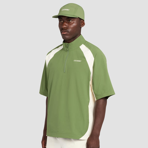 A man wearing a light green Reebok x Manors Men’s Tour Shirt with white side panels, paired with a matching green cap and white pants, stands against a plain gray background.