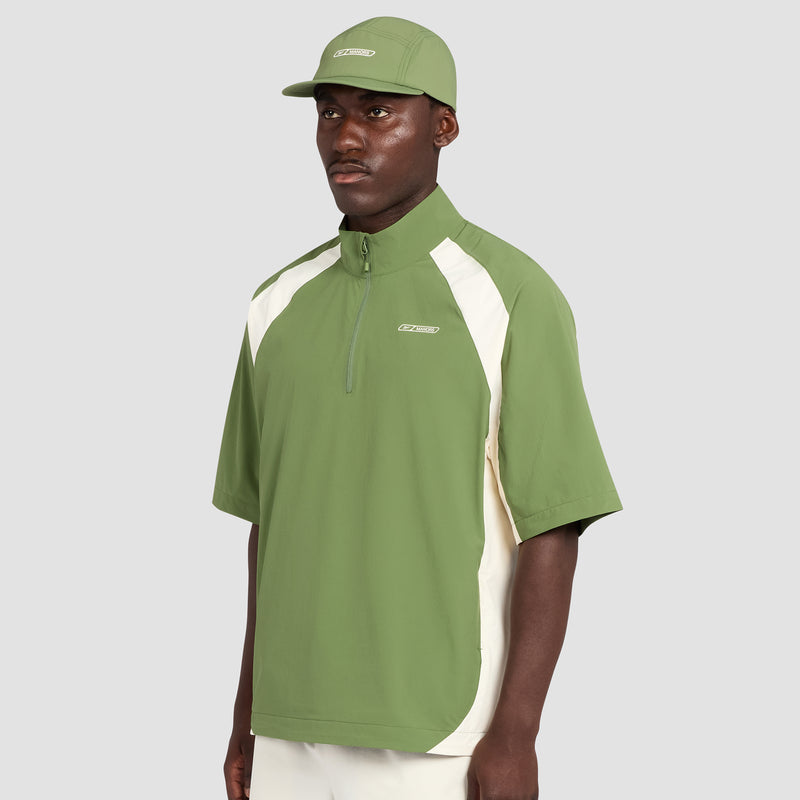 A man wearing a light green Reebok x Manors Men’s Tour Shirt with white side panels, paired with a matching green cap and white pants, stands against a plain gray background.
