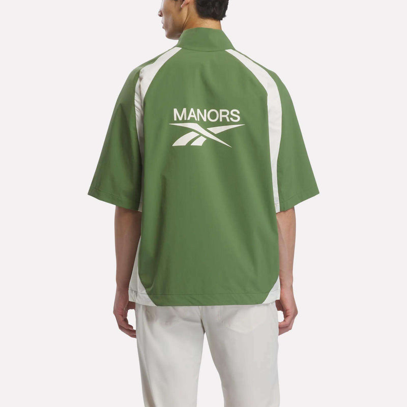 A person is shown from behind against a plain light background, wearing the Reebok x Manors Men’s Tour Shirt in green and white with short sleeves, featuring ‘MANORS’ and a logo on the back, paired with white pants.