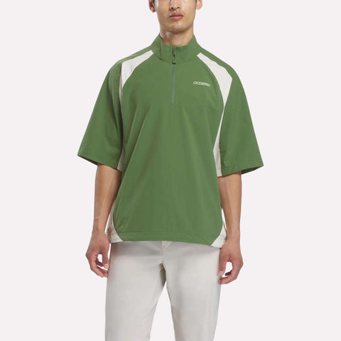 A man wearing the Reebok x Manors Men’s Tour Shirt by Reebok in green and white short-sleeves, paired with light-colored pants, stands against a plain white background.