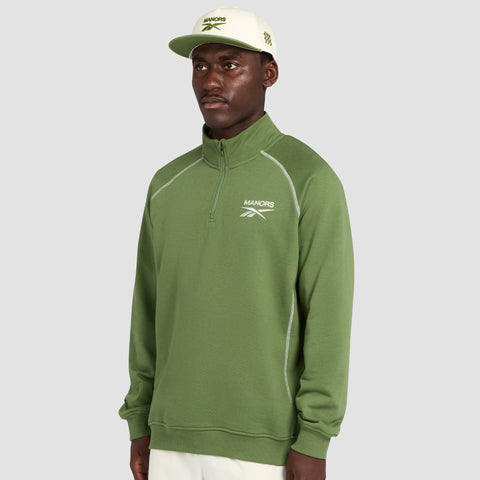 A man stands against a plain gray background wearing the Reebok x Manors Men’s Quarter-Zip Pullover in green, styled with white pants and a white cap.