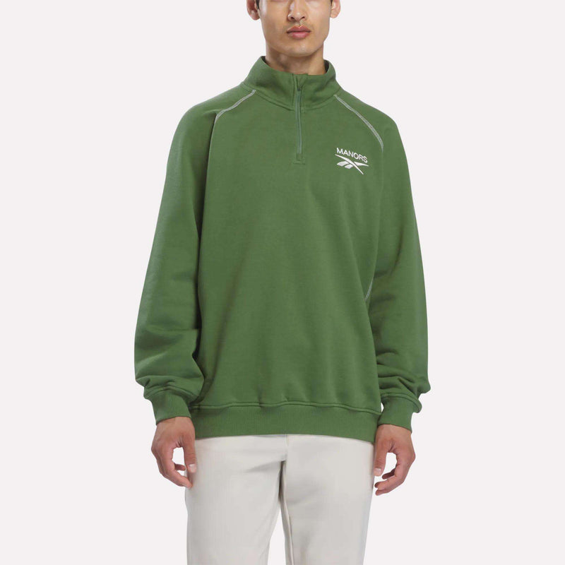 A person wearing the Reebok x Manors Men’s Quarter-Zip Pullover in green, with a white MANORS logo, paired with white pants, stands against a plain light background.