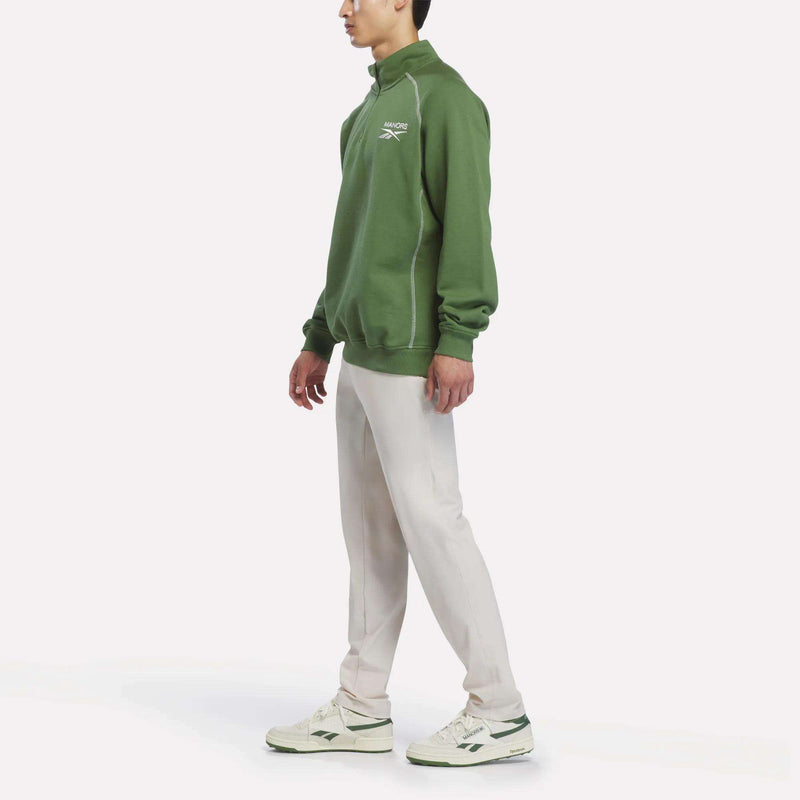 A person stands sideways against a plain white background, wearing the Reebok x Manors Men’s Quarter-Zip Pullover in green from Reebok, paired with light beige pants and white sneakers featuring green accents.
