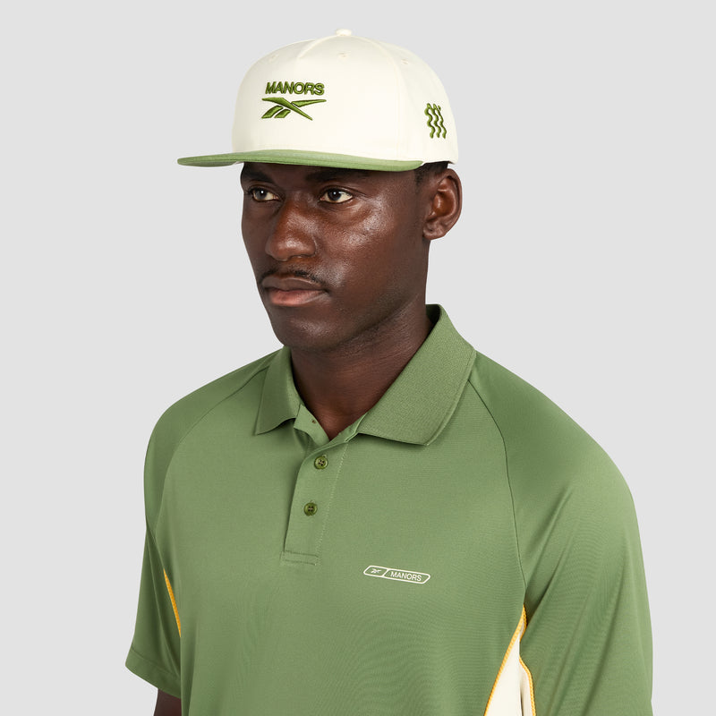 A man wearing the Reebok x Manors Men’s Retro Crown Cap in cream with green embroidery and a green collared polo shirt stands against a plain gray background, looking slightly to the side with a neutral expression. The retro crown design of his Reebok cap introduces a vintage flair to his understated ensemble.