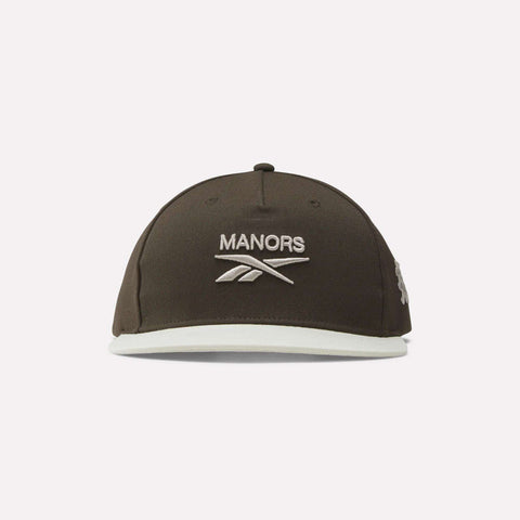 Introducing the Reebok x Manors Men’s Retro Crown Cap, a dark brown baseball cap featuring MANORS and the Reebok logo embroidered in white on the front. This cap showcases a retro crown silhouette, complemented by a light-colored brim, an adjustable snapback closure, and a small logo on the left side.