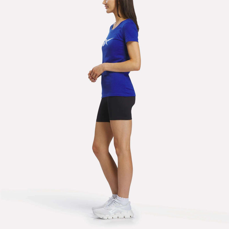 A woman stands sideways against a plain white background, wearing the Reebok Women’s Logo Tee in blue, paired with black shorts, white socks, and white sneakers.