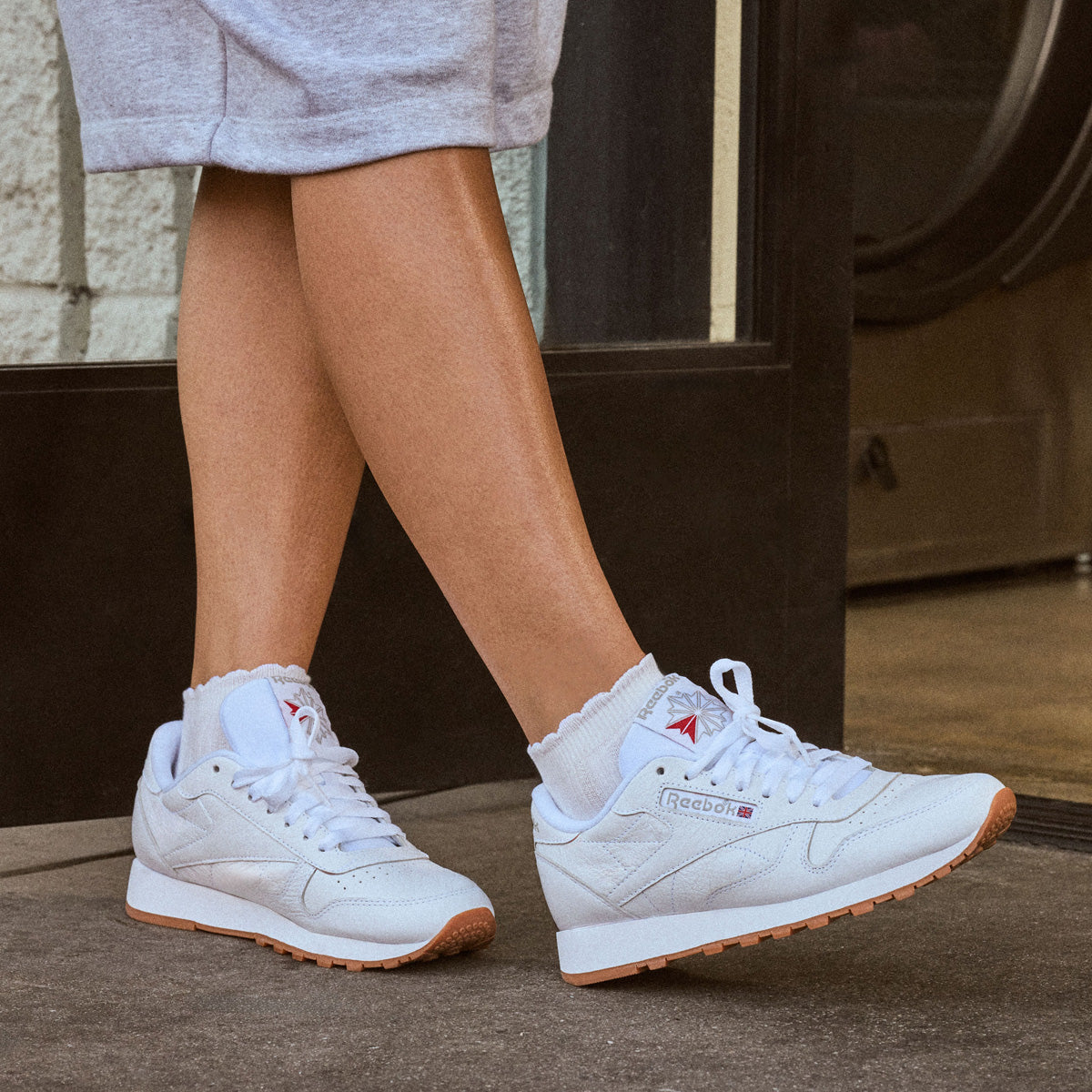 White Reebok sneakers with white socks and a light gray skirt, shown on a person's feet in an indoor setting.