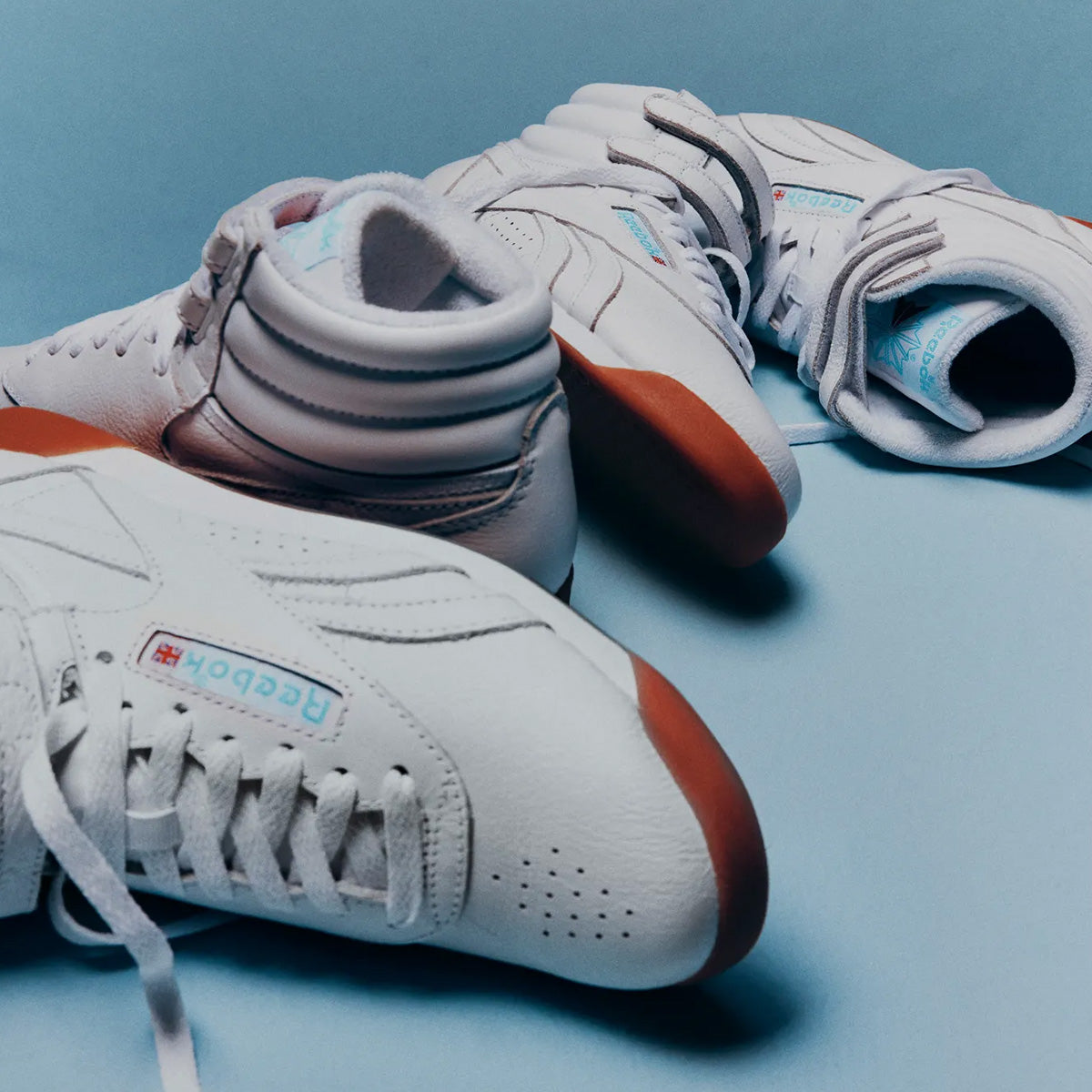 A pair of Reebok high-top sneakers in white with light blue accents and gum brown soles, arranged on a light blue background.