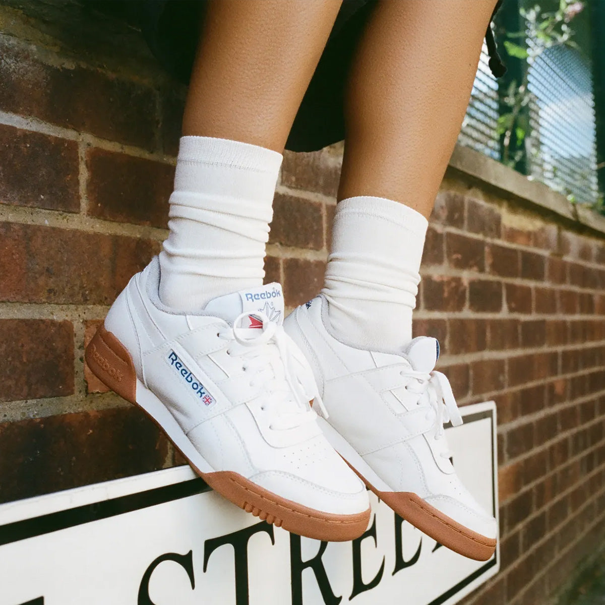 White Reebok sneakers with gum soles and white socks, worn by a person sitting against a brick wall.