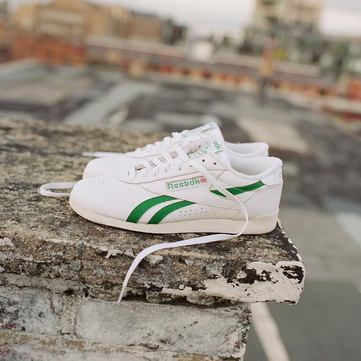 A pair of white Reebok sneakers with green stripes resting on a weathered concrete ledge with a blurred outdoor city background.