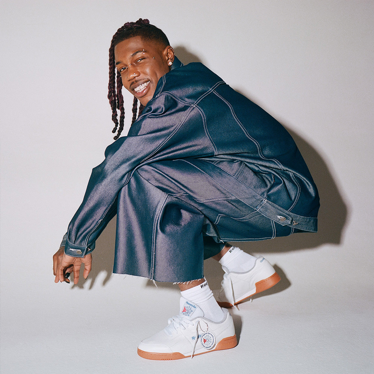 A man crouching in a studio setting, wearing a shiny dark blue denim jacket and matching denim pants with white sneakers featuring orange gum soles.