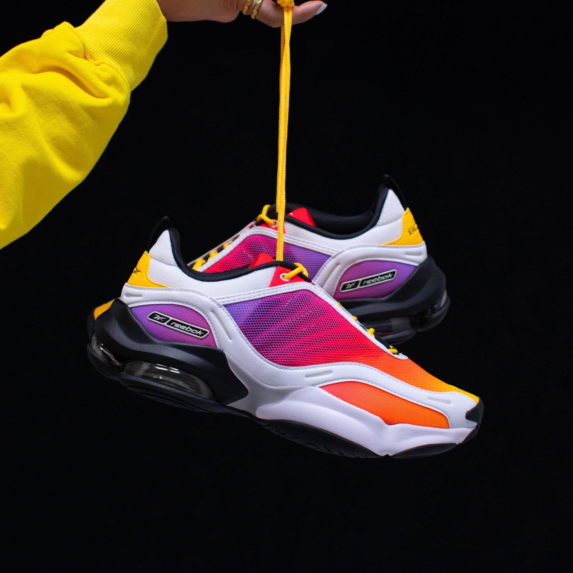 A hand in a yellow sleeve holds up the vibrant Reebok x Brain Child DMX Series 3000 Shoes by their yellow laces, featuring responsive cushioning and bold orange, purple, red, yellow, black, and white accents—part of the Red Nebula collab.