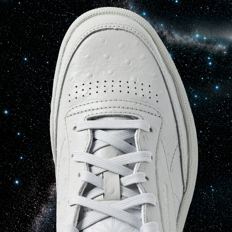 A close-up of a white Reebok x BBC Club C 85 shoe’s toe and laces appears against a starry outer space background, with a visible galaxy highlighting the iconic silhouette of these heritage Reebok sneakers.