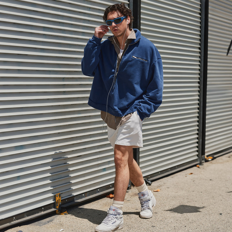 Person wearing a blue jacket and white sneakers standing against a corrugated metal wall.