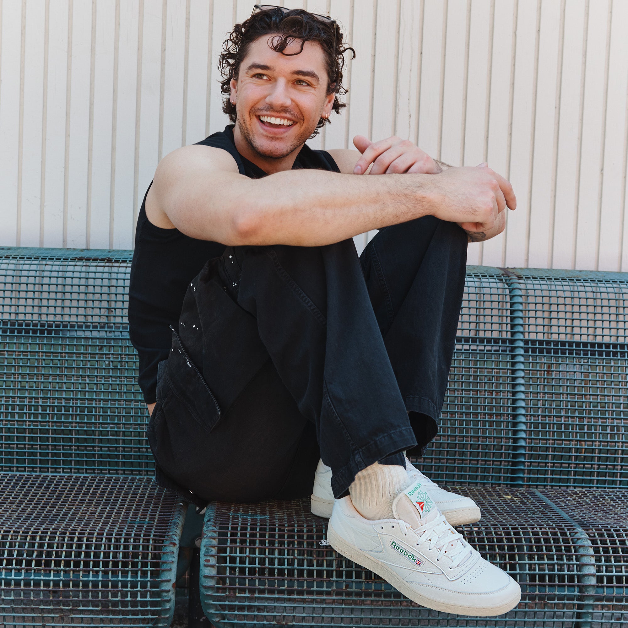 Man sitting on a metal bench wearing a black jacket, dark pants, and white sneakers.