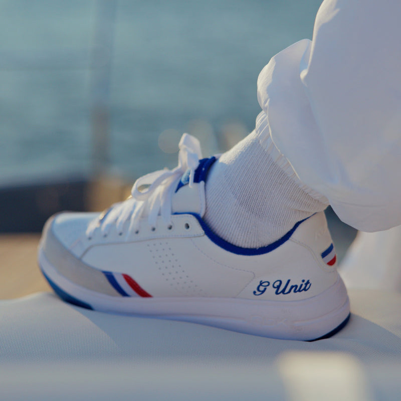A close-up of a person wearing the Reebok x G-Unit G6 Men’s Shoes in white with blue trim, showcasing hip-hop sneaker style and the signature G Unit branding on the side. The mix of leather and suede materials is highlighted, paired with white pants. The background is blurred, featuring hints of water and wood.