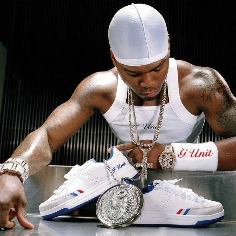 A man in a white tank top, durag, and G Unit wristband leans over a table with hip-hop sneakers and a large G medallion necklace. He is heavily tattooed and wearing a cross necklace, a luxury watch, and white Reebok x G-Unit G6 Men’s Shoes from Reebok crafted from leather and suede.