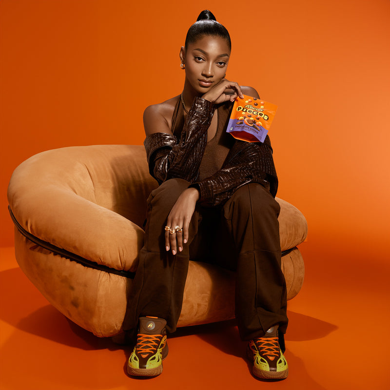 A woman sits on a tan chair against an orange background, holding a bag of TCHO Choco Popped snacks. She wears a dark outfit and Reese’s x Angel Reese 1 Basketball Shoes by Reebok, looking confidently at the camera.