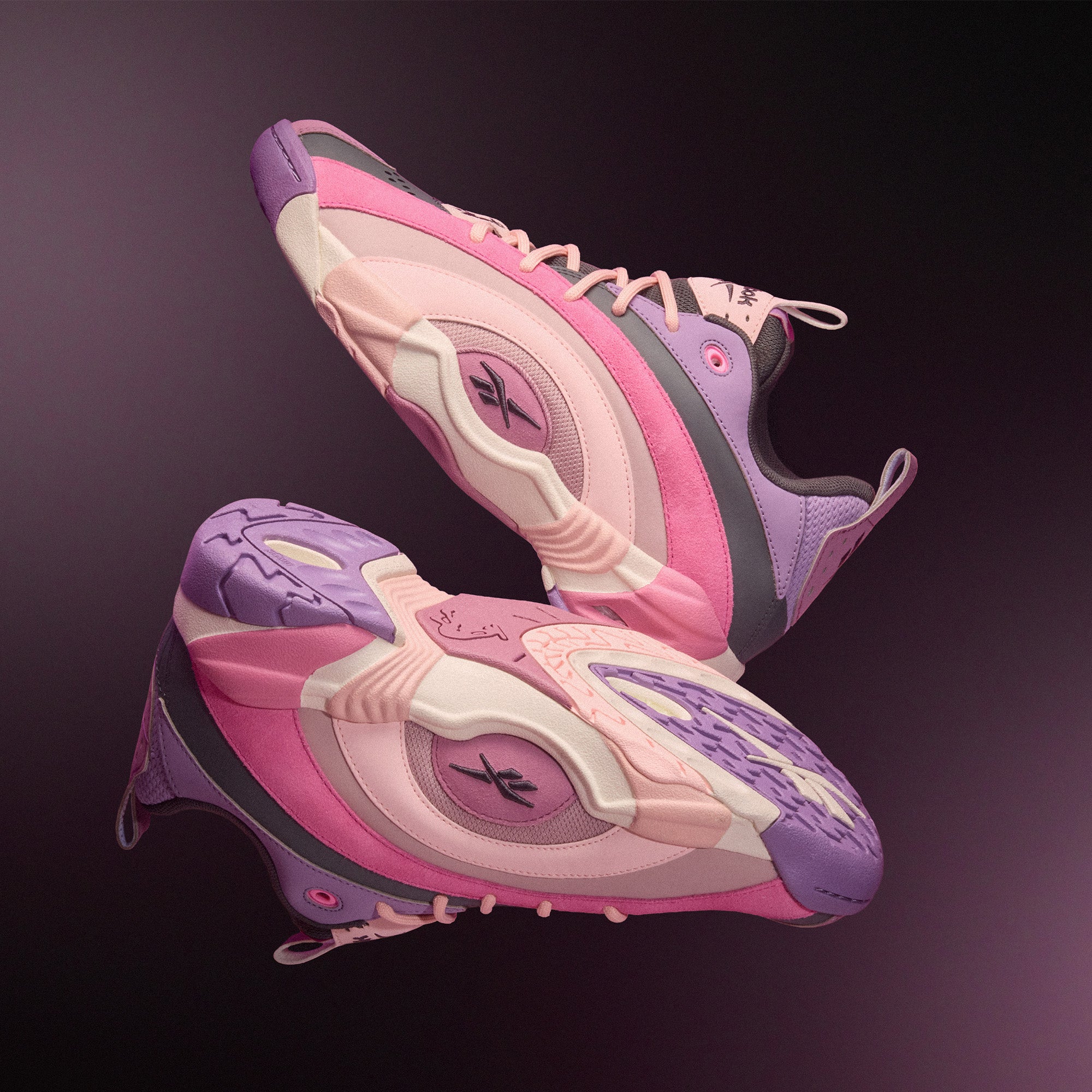 Two pink and purple Reebok Shaqnosis Low Shoes hover against a dark gradient backdrop, displaying side and sole views with stylized patterns, logo accents, and visible Hexalite cushioning.