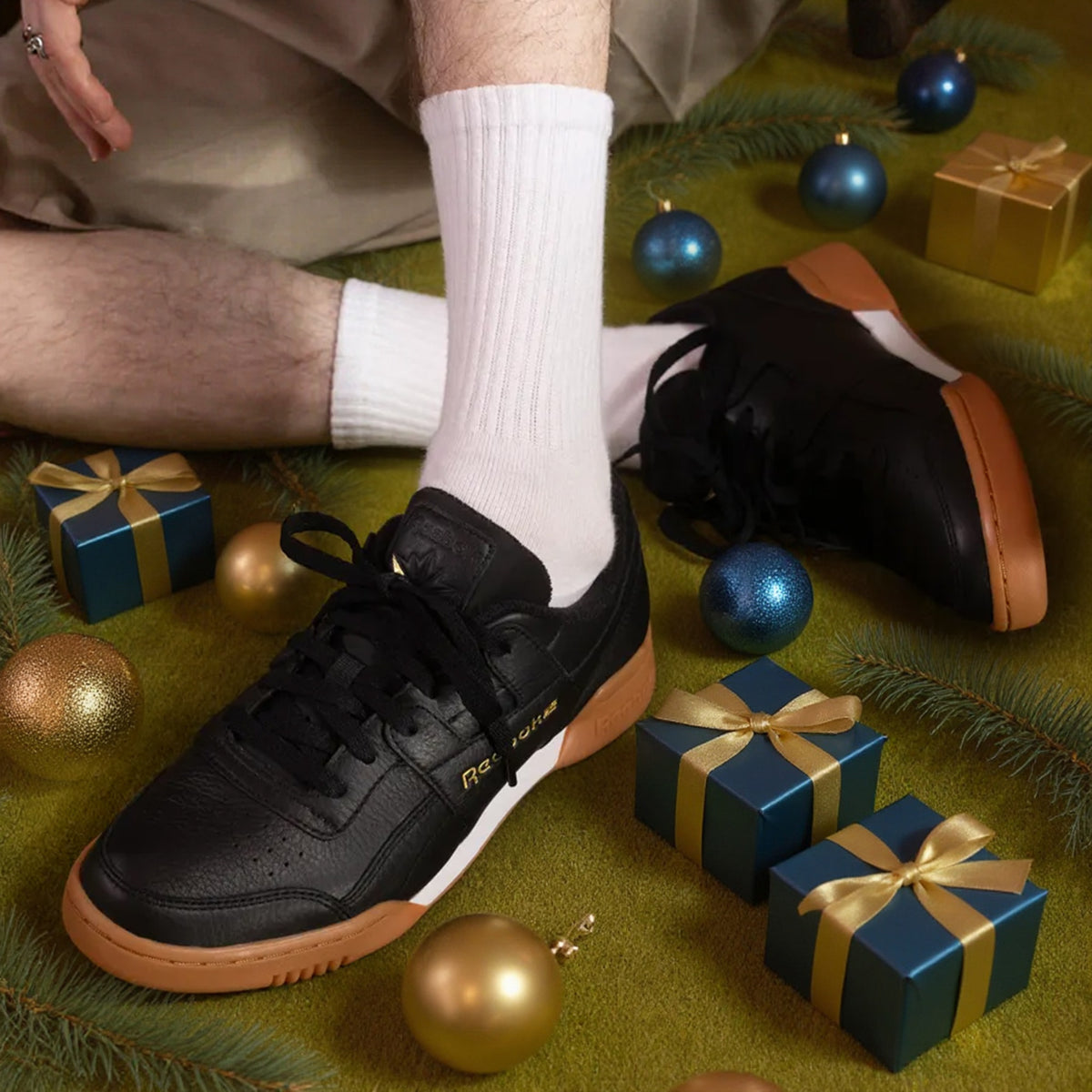 A person wearing black Reebok sneakers with white socks, seated on a green festive surface decorated with Christmas ornaments and gift boxes. The shoes feature black leather uppers, black laces, a white midsole stripe, and gum brown soles.
