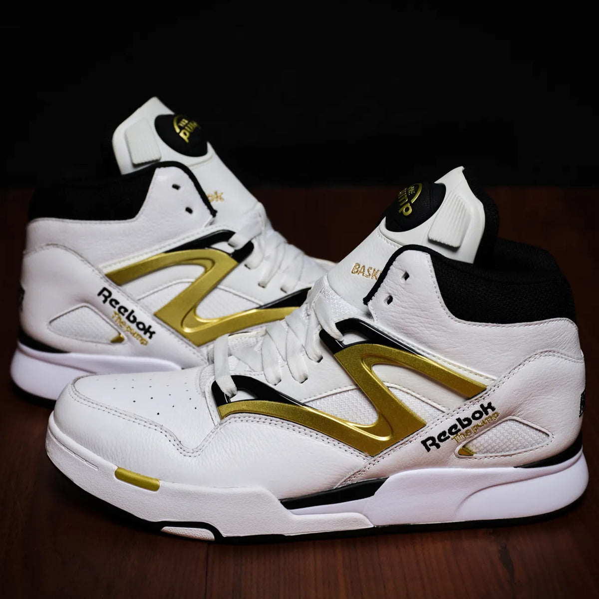 A pair of Reebok high-top basketball sneakers in white leather with prominent metallic gold and black accents, displayed on a dark wooden surface against a black background.