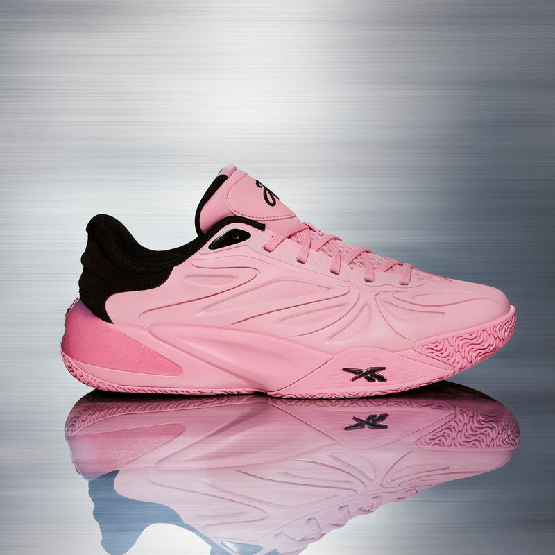 The Angel Reese 1 Basketball Shoes - Big Kids by Reebok are pink athletic sneakers with black accents and laces, displayed in profile on a reflective surface against a brushed metal background. Featuring the iconic Reebok logo on the side and tongue, these stylish kids’ basketball shoes are perfect for young athletes who want to stand out—just like Angel Reese.