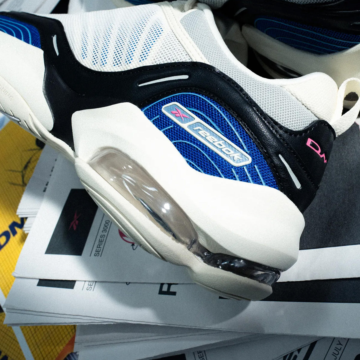 A close-up of a Reebok sneaker with a white, black, and blue colorway, featuring a prominent clear air cushion unit in the midsole, resting on a stack of papers.