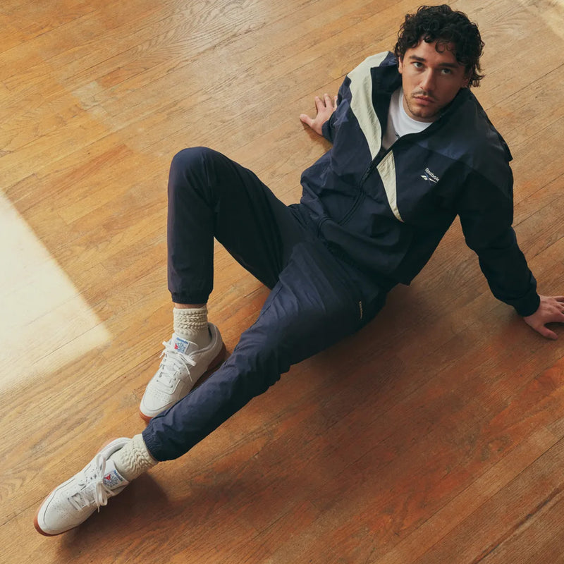 Dressed in a navy tracksuit and white Reebok Workout Plus Shoes, Belmont Cameli channels classic ’80s fitness style as he sits on a wooden floor, leaning back on one arm with one leg bent and the other extended. He gazes up at the camera with a serious expression, his Reebok Workout Plus Shoes completing his retro-inspired look.