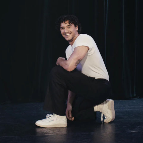Belmont Cameli wearing a white t-shirt, black pants, and Reebok Club C 85 Vintage Shoes featuring lightweight cushioning and a leather upper, kneels on one knee and smiles in front of a dark curtain.