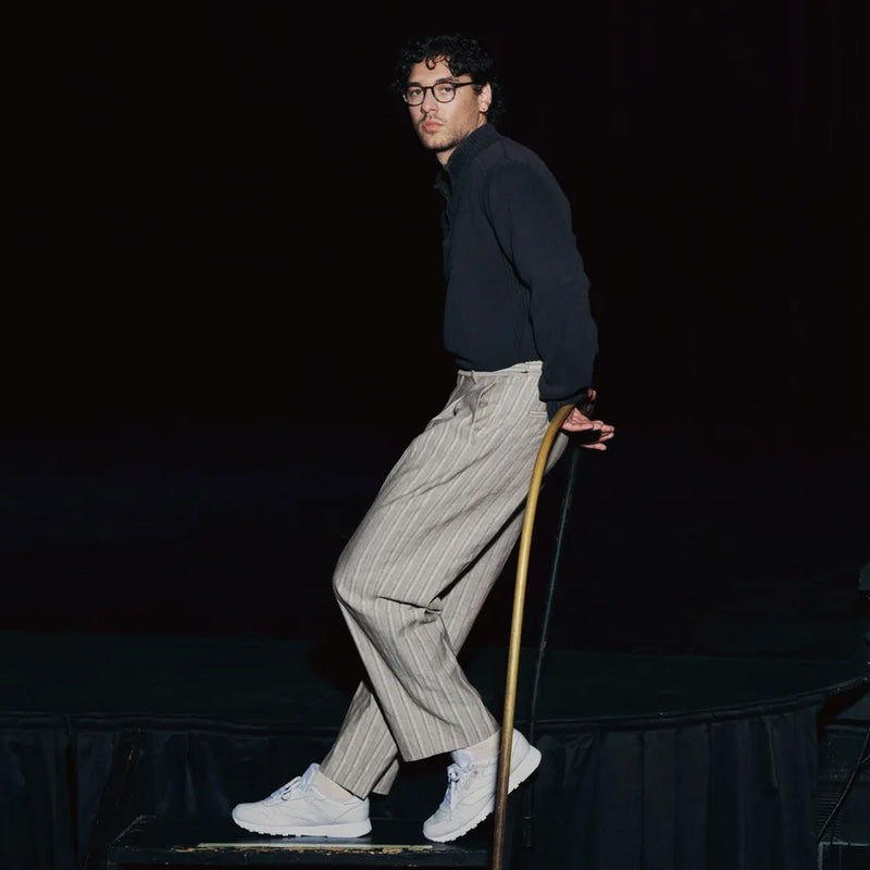 Belmont Cameli wearing glasses, dressed in a dark shirt, striped pants, and white sneakers— the Classic Leather Shoes by Reebok—sits on a railing against a dark background, highlighting their retro footwear.