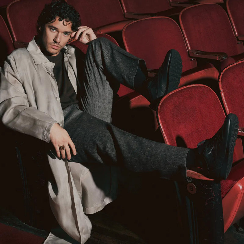 Belmont Cameli wears a light trench coat sits casually in a red theater seat, with one leg draped over the armrest and their other foot resting on the seat ahead. They wear Reebok Classic Leather Shoes and gaze at the camera with a relaxed expression.