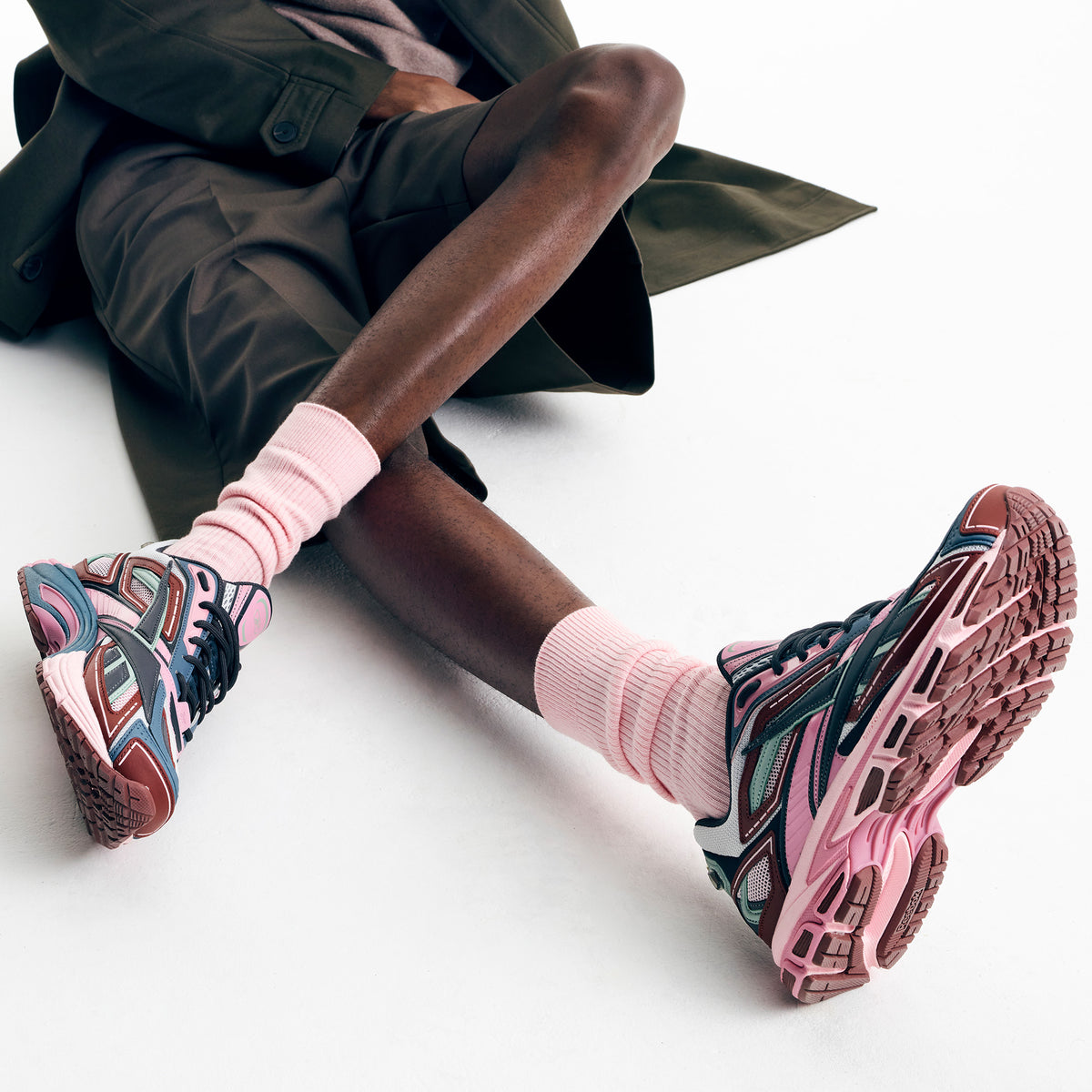 A person reclining on a white floor wearing chunky multicolor sneakers with pink laces and soles, pink crew socks, and an olive green coat draped over them.
