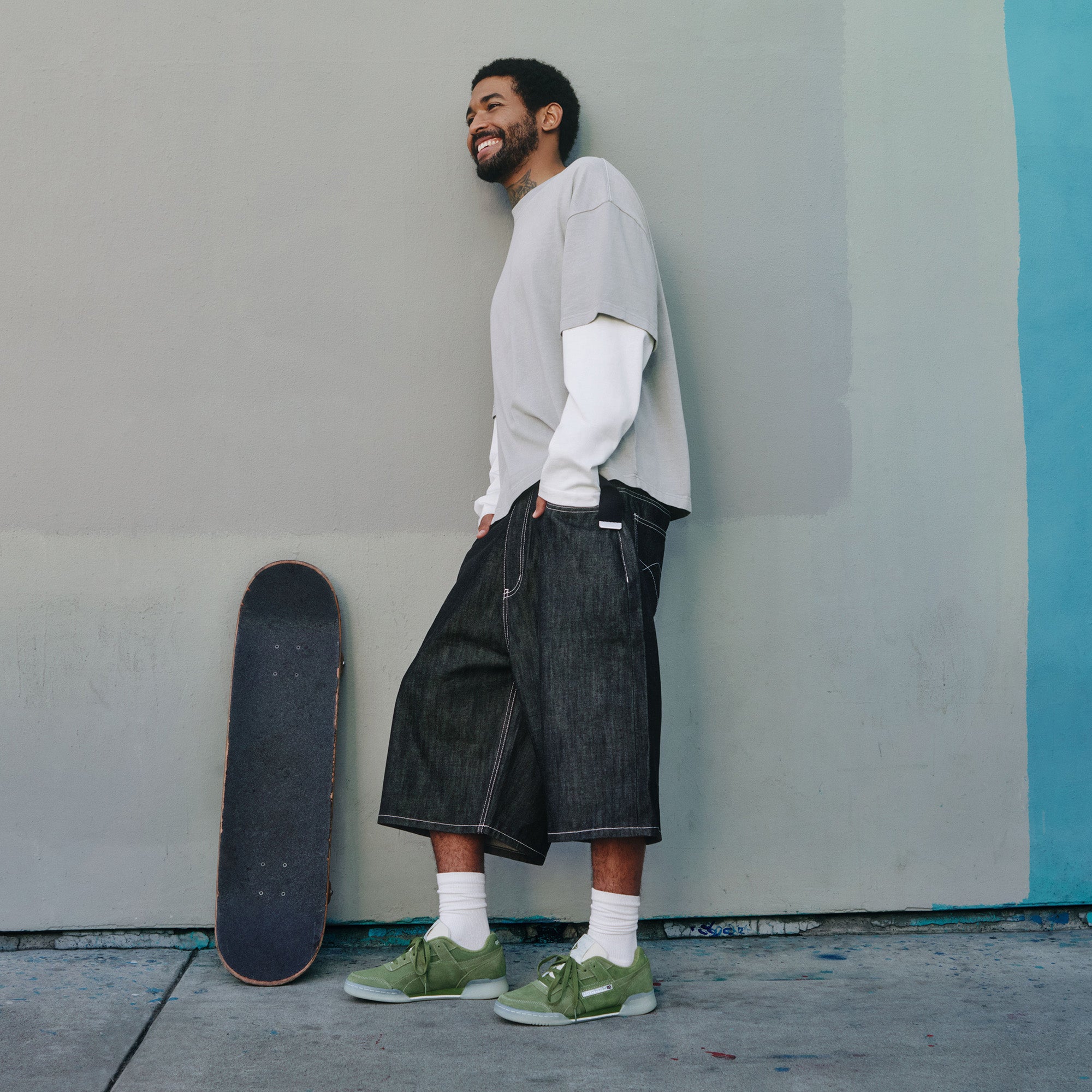 A man with a beard stands smiling against a light gray wall, wearing a beige t-shirt layered over a white long-sleeve shirt, baggy dark denim shorts, white socks, and green sneakers. A skateboard rests vertically nearby.