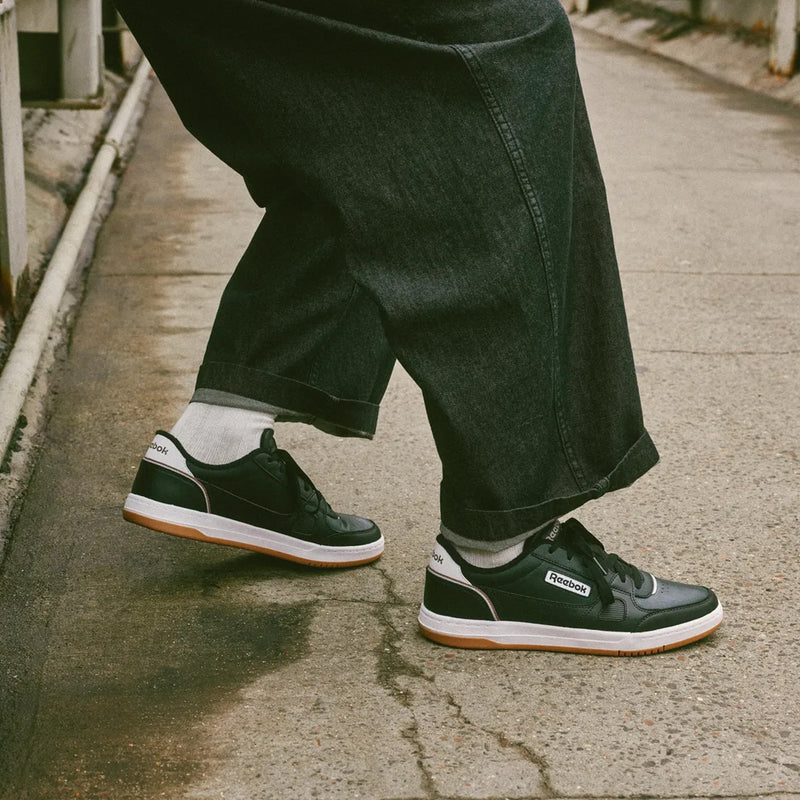 A person stands on a sidewalk, wearing black Reebok Men’s Court LXS Shoes with white soles and white socks. Only their lower legs and the wide-legged dark jeans are visible. These Reebok shoes are crafted from premium full grain leather, offering a classic and durable style.