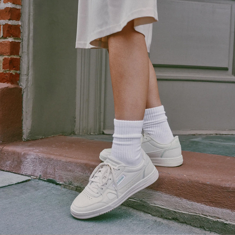 A person wearing off-white low-top sneakers with white crew socks, standing on a brick doorstep in front of a gray door.