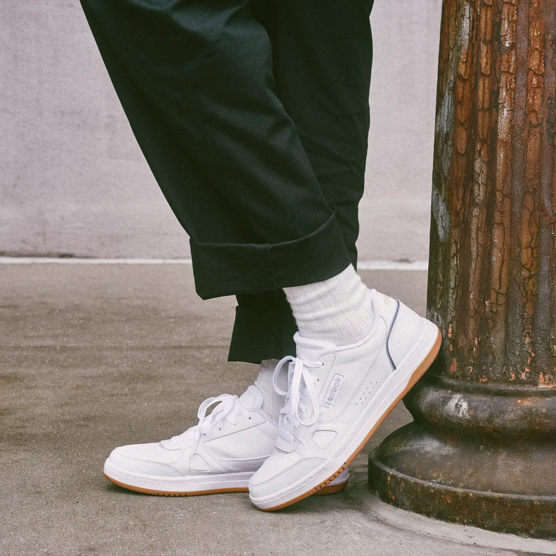 A person wearing crisp white Reebok Men’s Court LXS Shoes and white socks, paired with black pants, leans against a rusty metal pole on a concrete sidewalk. The background is a plain light-colored wall.