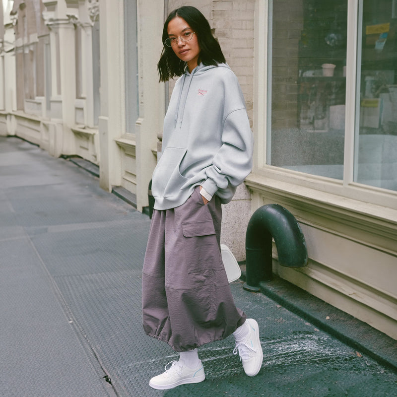 A woman stands on a city sidewalk wearing a light gray oversized hoodie, loose mauve cargo pants, white socks, and white sneakers, with storefront windows and a building facade in the background.