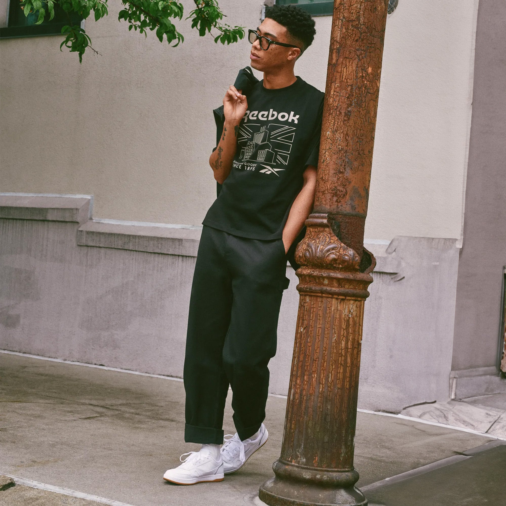 A man wearing a black Reebok sleeveless graphic T-shirt, black cargo pants, and white sneakers leans against a weathered wooden pole on a city sidewalk.