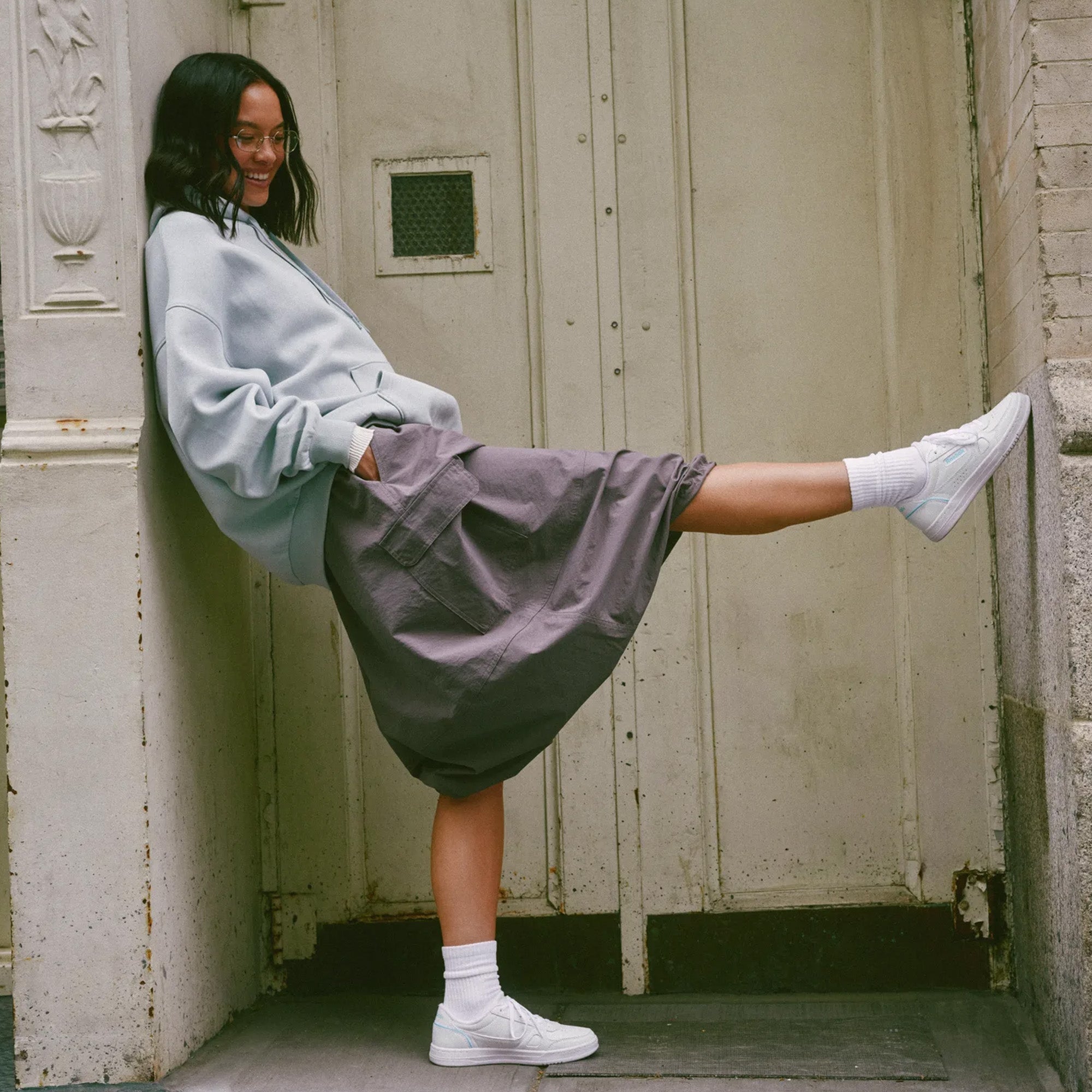 A woman leans against a weathered beige door, wearing a light blue oversized sweatshirt, a dark gray skirt, white socks, and white sneakers.