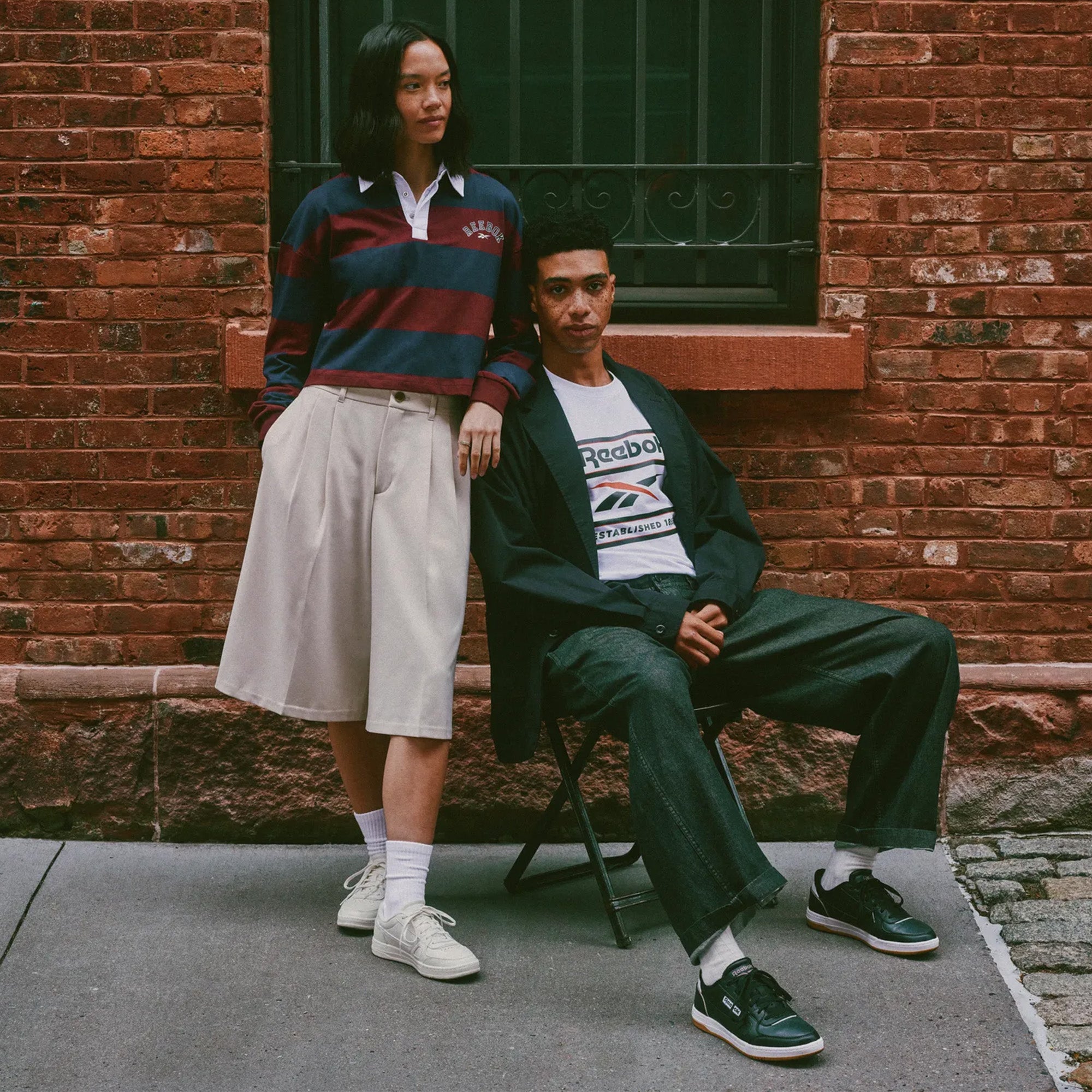Two models posing outdoors against a red brick wall. The standing model wears a navy and burgundy striped long-sleeve polo with a cream pleated skirt, white socks, and Reebok sneakers.