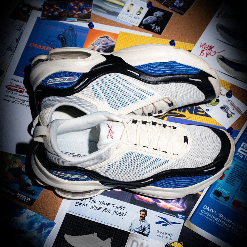 A pair of Reebok sneakers in white, blue, and black, styled on top of a corkboard covered with various printed magazine and poster pages.