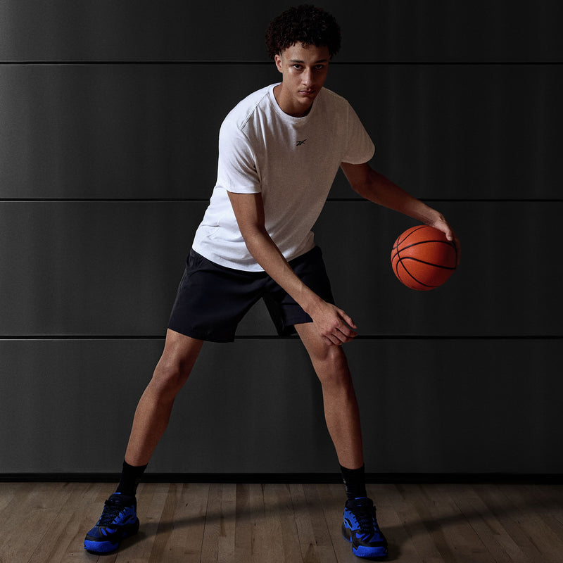 A young man in a white t-shirt, black shorts, and Reebok Engine A 26 Basketball Shoes dribbles a basketball indoors on a wooden court, highlighting energy return and stability against a sleek, modern dark wall.