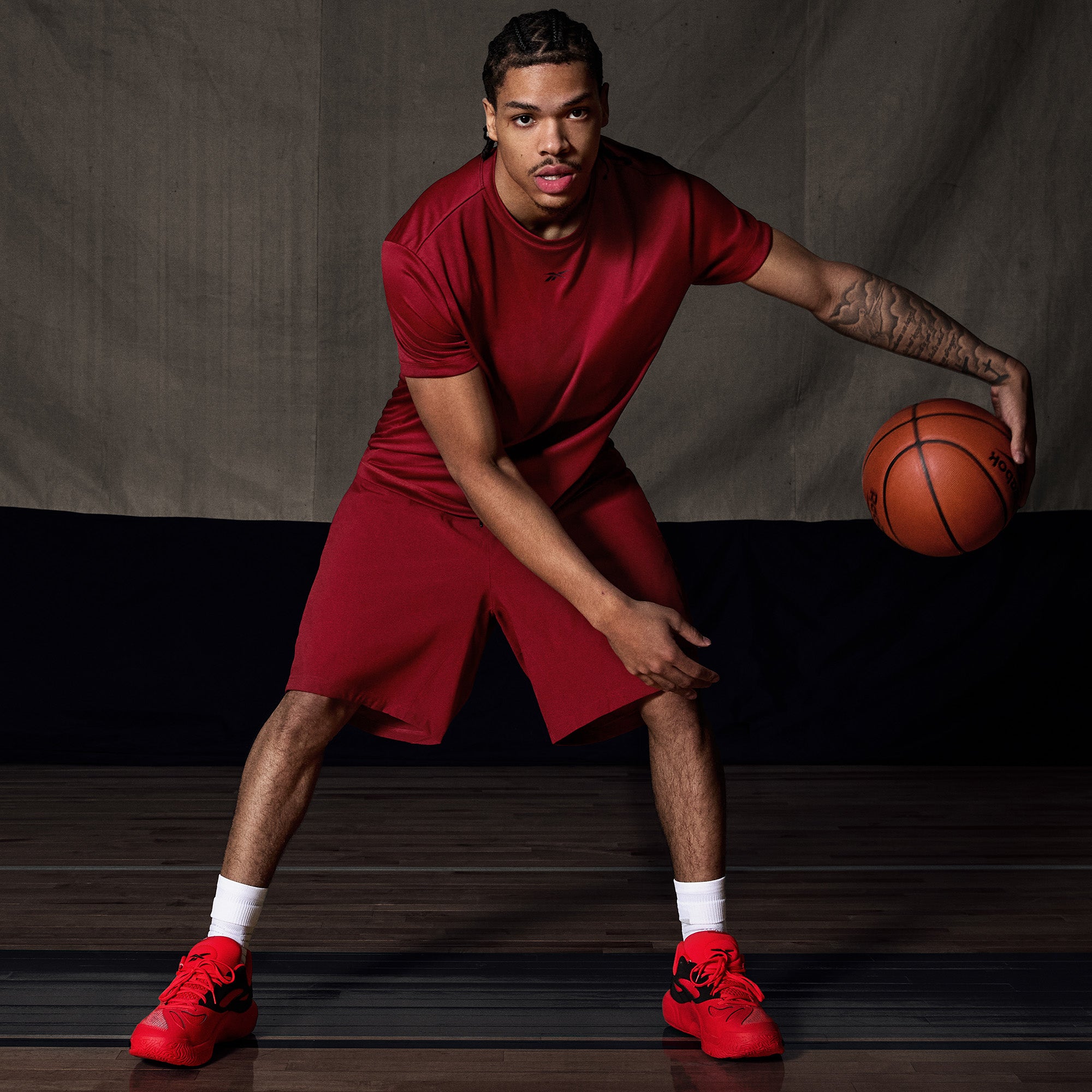A man in a red outfit and Reebok Engine A 26 Basketball Shoes dribbles on an indoor court, looking focused and poised, showcasing the shoes’ energy return and stability against a dark background.